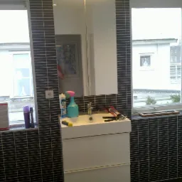 Wykończona łazienka z ciemną mozaiką, białą szafką z umywalką i oknem z widokiem na budynek. Widoczne narzędzia i preparaty, sugerujące świeży remont.