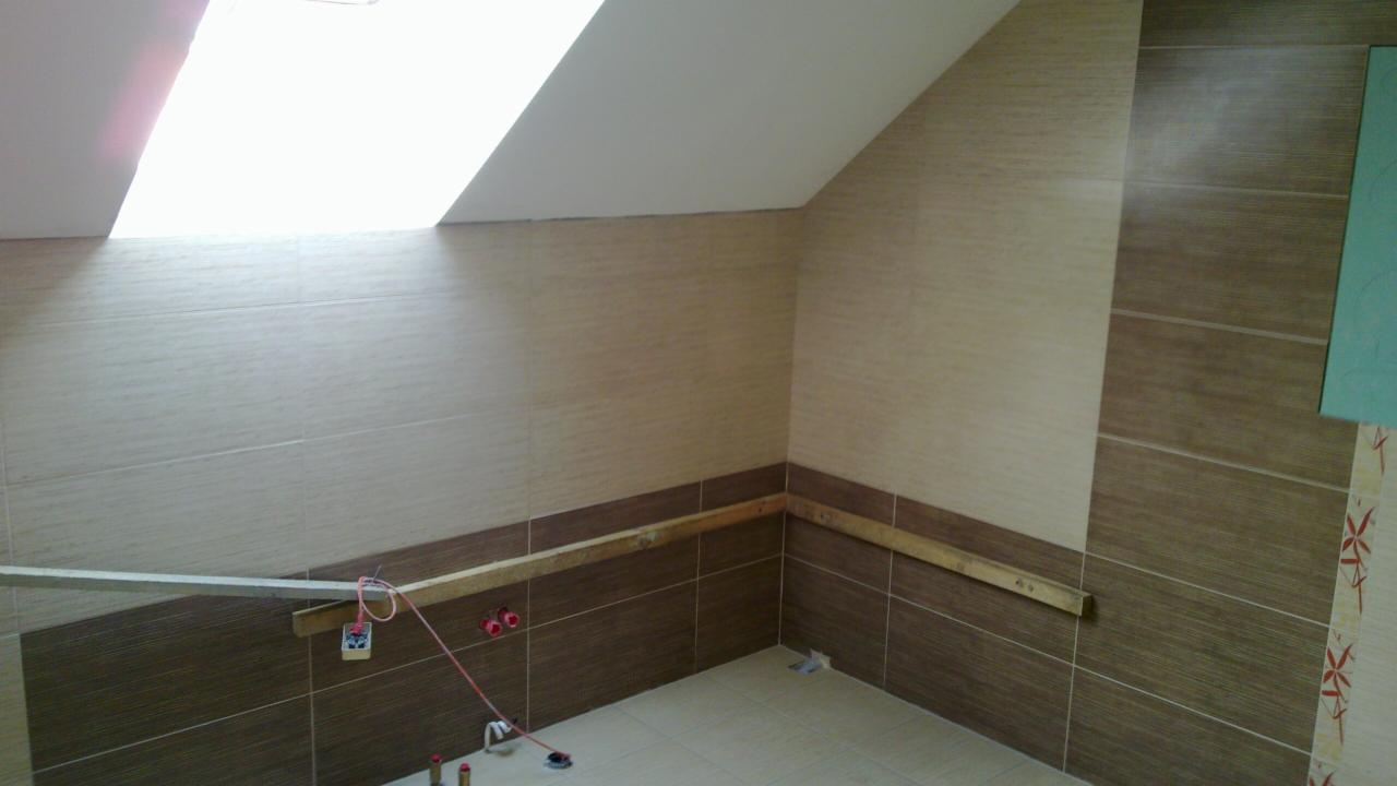 Układanie płytek w łazience na poddaszu z oknem dachowym. Widoczne brązowe i beżowe płytki, poziomica i narzędzia glazurnicze. Wnętrze w trakcie wykańczania.