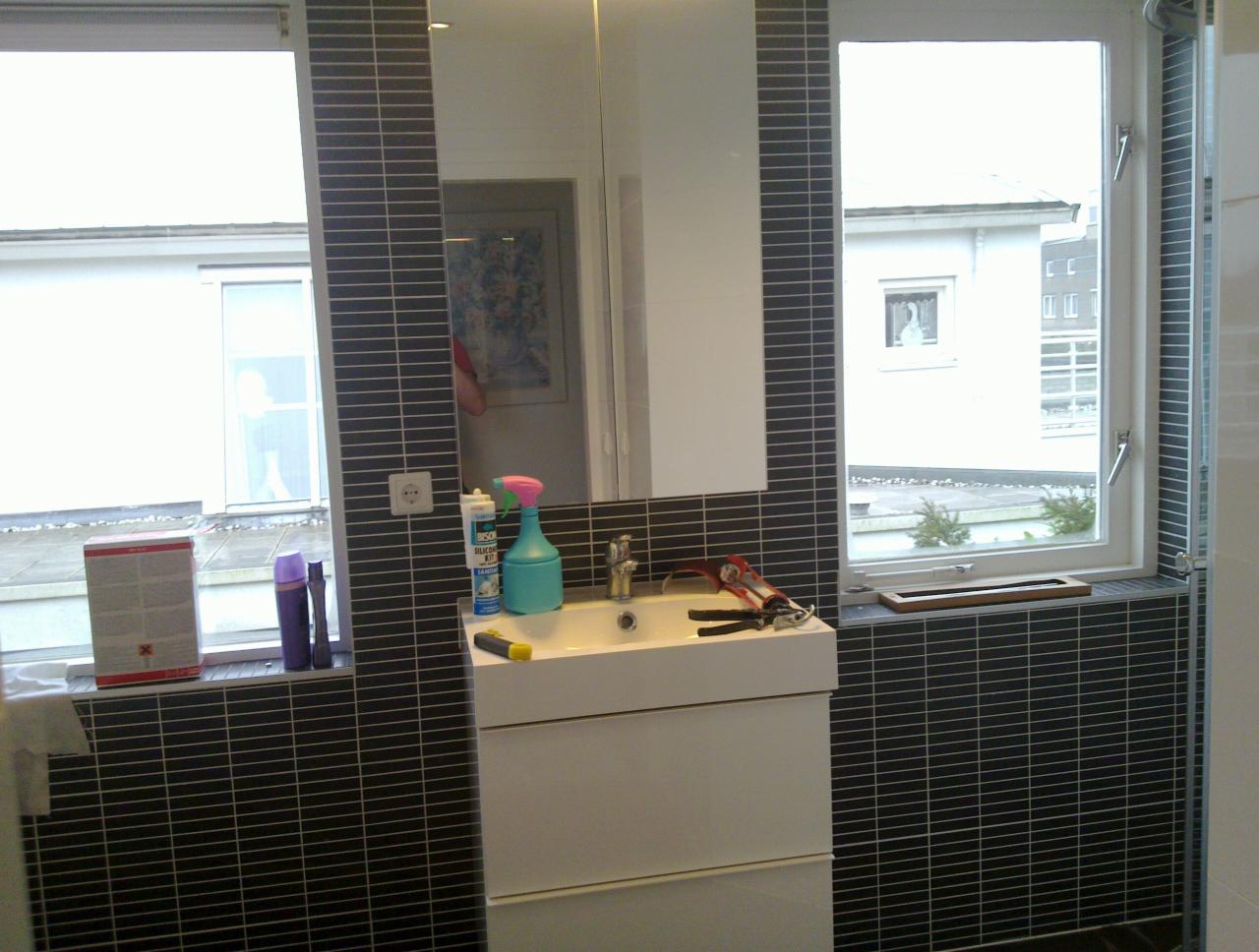Wykończona łazienka z ciemną mozaiką, białą szafką z umywalką i oknem z widokiem na budynek. Widoczne narzędzia i preparaty, sugerujące świeży remont.