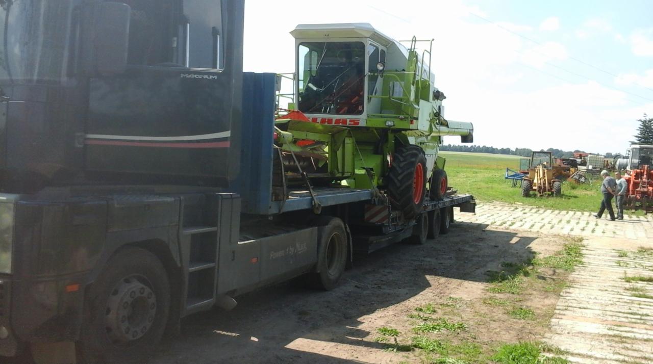 Ciężarówka Magnum transportuje kombajn Claas na niskopodwoziowej lawecie. Widoczne pole i inne maszyny rolnicze w tle. Dwóch mężczyzn stoi obok drogi z płyt.