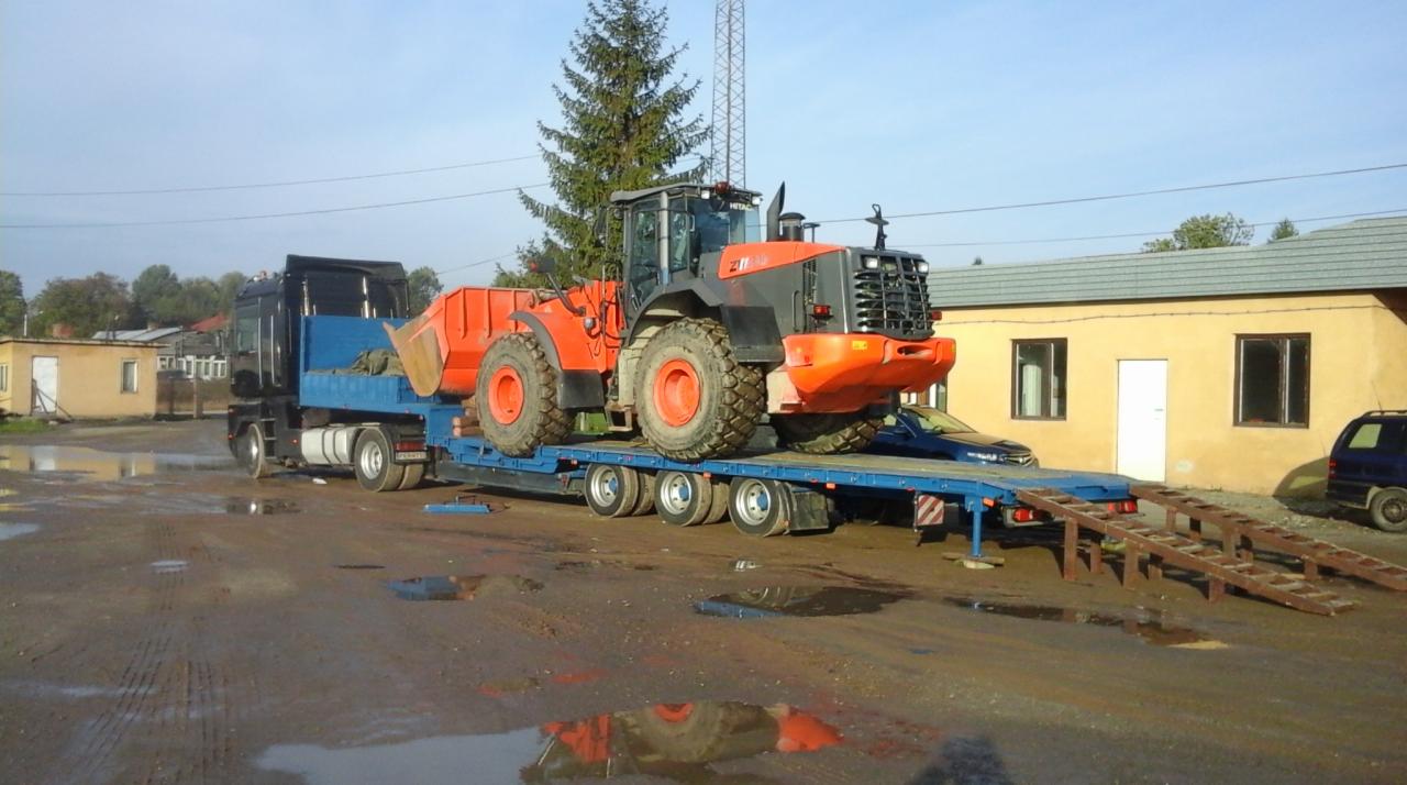 Pomarańczowa ładowarka kołowa na niebieskiej lawecie z najazdami, transportowana przez czarny ciągnik siodłowy, na placu z kałużami.