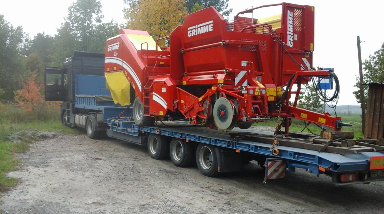 Czerwony kombajn ziemniaczany marki Grimme przewożony na niebieskiej niskopodwoziowej lawecie, ciągniętej przez czarny ciągnik siodłowy. Tło stanowią drzewa.