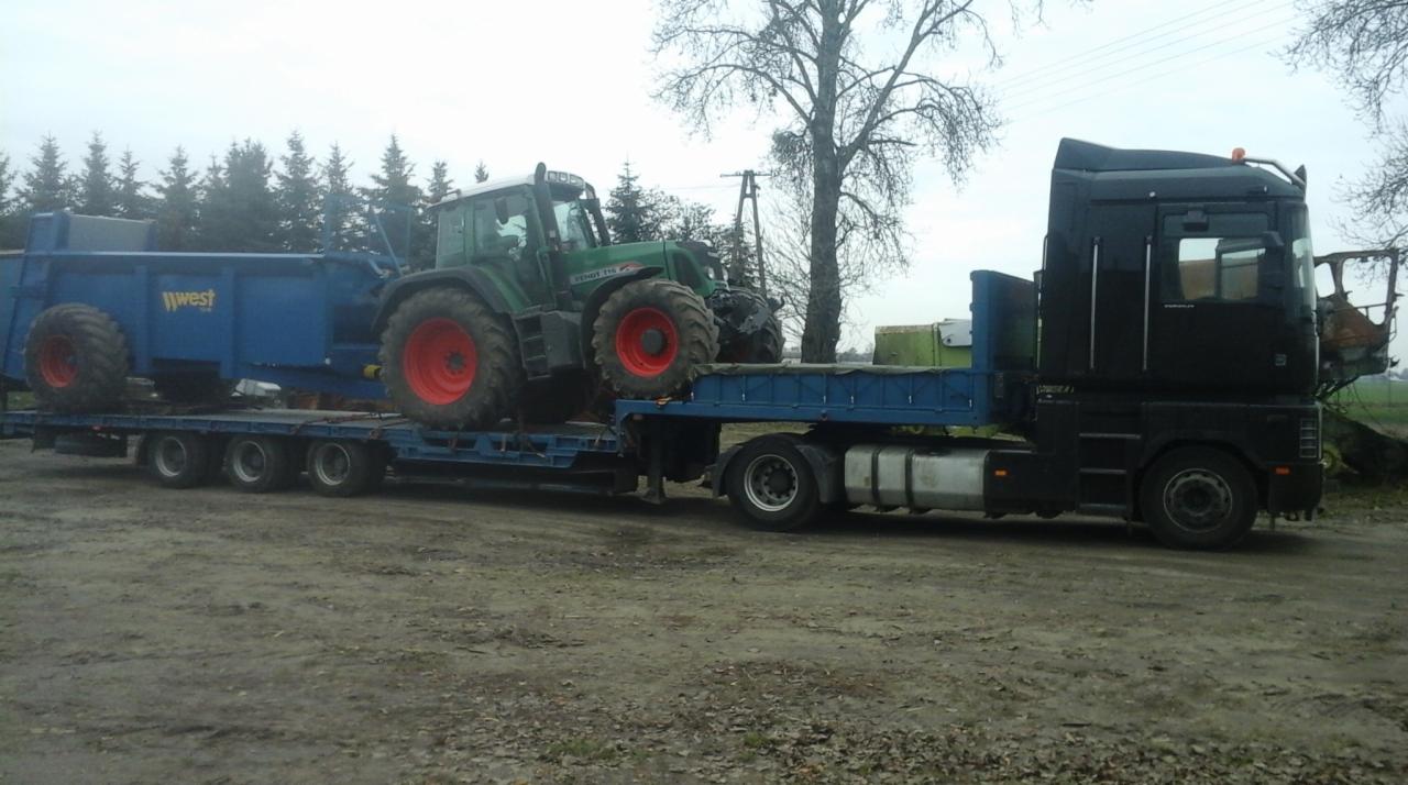 Czarna ciężarówka transportuje na lawecie zielony traktor Fendt i niebieską przyczepę rolniczą marki West na tle drzew i pochmurnego nieba.