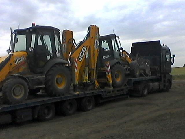 Laweta przewozi dwie żółto-czarne koparko-ładowarki JCB. Jedna z nich jest częściowo schowana za drugą. Sceneria wiejska, pochmurne niebo.