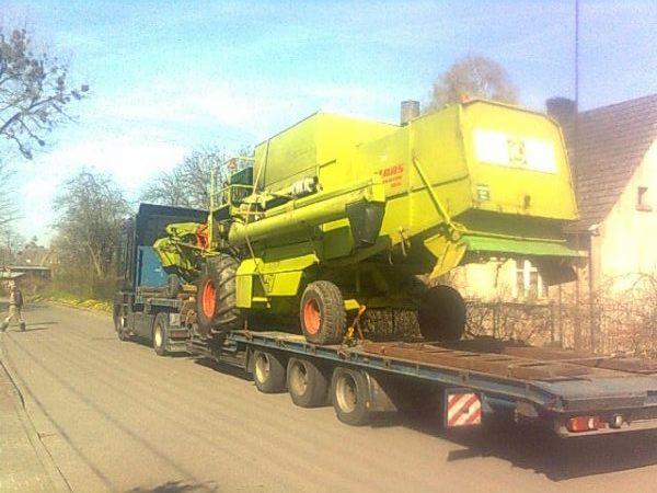Transport kombajnu CLAAS na niskopodwoziowej platformie. Widoczny fragment budynku w tle i osoba przechodząca obok. Jasne niebo.