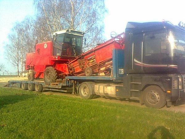 Czerwony kombajn na niskopodwoziowej naczepie ciągniętej przez czarny ciągnik siodłowy. Transport maszyny rolniczej na tle zielonej trawy i drzew.