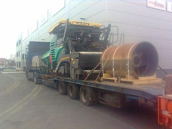 Laweta przewozi walec drogowy i maszynę do asfaltu marki Vogele. Transport ciężarowy na platformie, zabezpieczony pasami, na tle szarego budynku.