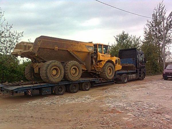 Żółty, brudny wywrotka Volvo na niskopodwoziowej naczepie ciężarówki, transportowana na placu budowy. Widok z boku, pochmurne niebo.