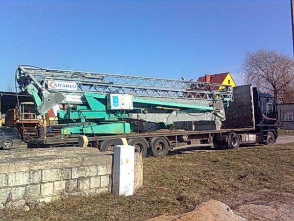 Ciężarówka przewozi złożony, zielony dźwig Cattaneo na platformie. Widoczne elementy konstrukcyjne i logo producenta. Na drugim planie budynek z żółtą ścianą.