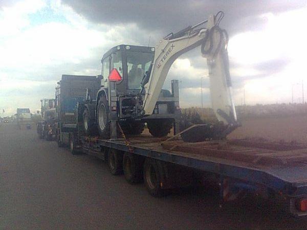 Laweta przewozi koparkę Terex na niskopodwoziowej platformie. Widok z boku, w tle inne pojazdy i pochmurne niebo.