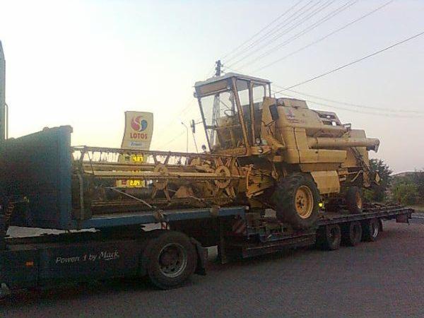 Żółty kombajn zbożowy na niskopodwoziowej lawecie, transportowany w pobliżu stacji benzynowej Lotos. Widok z boku na drogę i niebo.