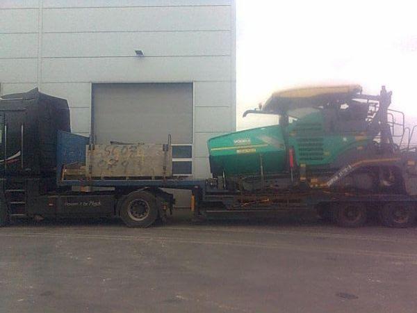 Transport niskopodwoziowy: ciężarówka z platformą przewozi zielony rozściełacz asfaltu na tle hali magazynowej. Widoczny element ładunku z numerem.