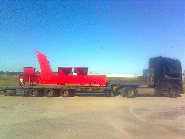 Czerwona konstrukcja na niskopodwoziowej naczepie transportowana ciężarówką na tle błękitnego nieba i zielonego terenu.