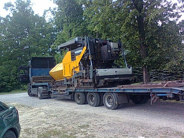 Żółty rozściełacz asfaltu marki Volvo przewożony na niskopodwoziowej lawecie, widoczny na żwirowej drodze w otoczeniu drzew.