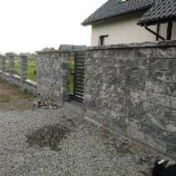 K&M - Fragment ogrodzenia z szarej, strukturalnej elewacji kamiennej z metalową furtką, widok z zewnątrz na żwirowy podjazd i dom w tle.