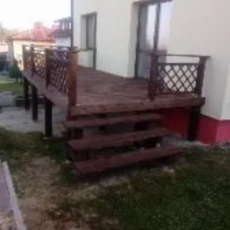 Drewniany taras przylegający do jasnej ściany domu z widocznymi schodami i balustradą z kratką, otoczony trawnikiem.