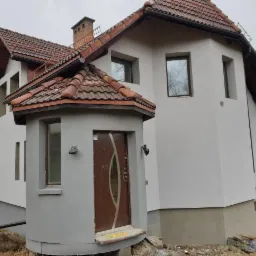 Dom jednorodzinny po nałożeniu jasnoszarego tynku zewnętrznego, widoczne okna z refleksami drzew i brązowe drzwi wejściowe z daszkiem krytym czerwoną dachówką.