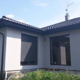 K&M - Jasnoszara elewacja domu jednorodzinnego z zamkniętą roletą zewnętrzną i dużym oknem, widoczne czarne rynny i dachówka oraz fragment palety pod oknem.