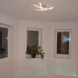 Wnętrze pokoju po tynkowaniu ścian, widoczne trzy okna, roślina doniczkowa na parapecie środkowego okna, nowoczesna lampa sufitowa, ślady gładzi szpachlowej na ścianach.