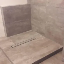 Wykończony brodzik z odpływem liniowym, obłożony płytkami imitującymi beton, z fragmentem ściany wykończonej białymi i szarymi płytkami.