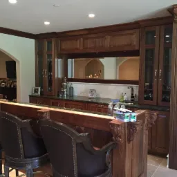 Zabudowa baru w stylu klasycznym z ciemnego drewna, z krzesłami barowymi z brązowej skóry i metalu, widoczne szklanki, butelki i dekoracje, marmurowy blat i lustro w tle.