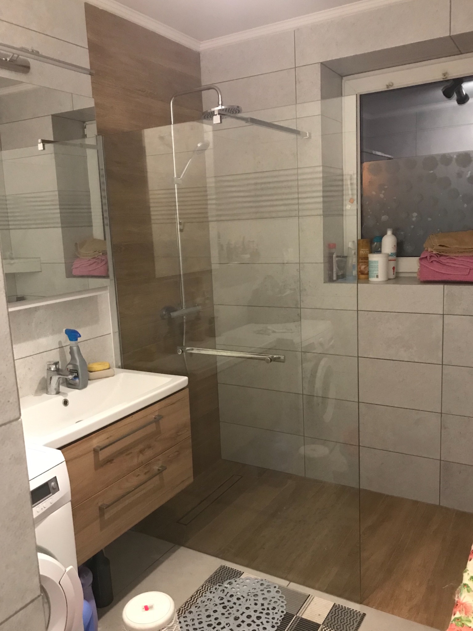 Nowoczesna łazienka z kabiną prysznicową typu walk-in, drewnianą zabudową umywalki i szarą płytką ścienną, widoczna pralka z boku.