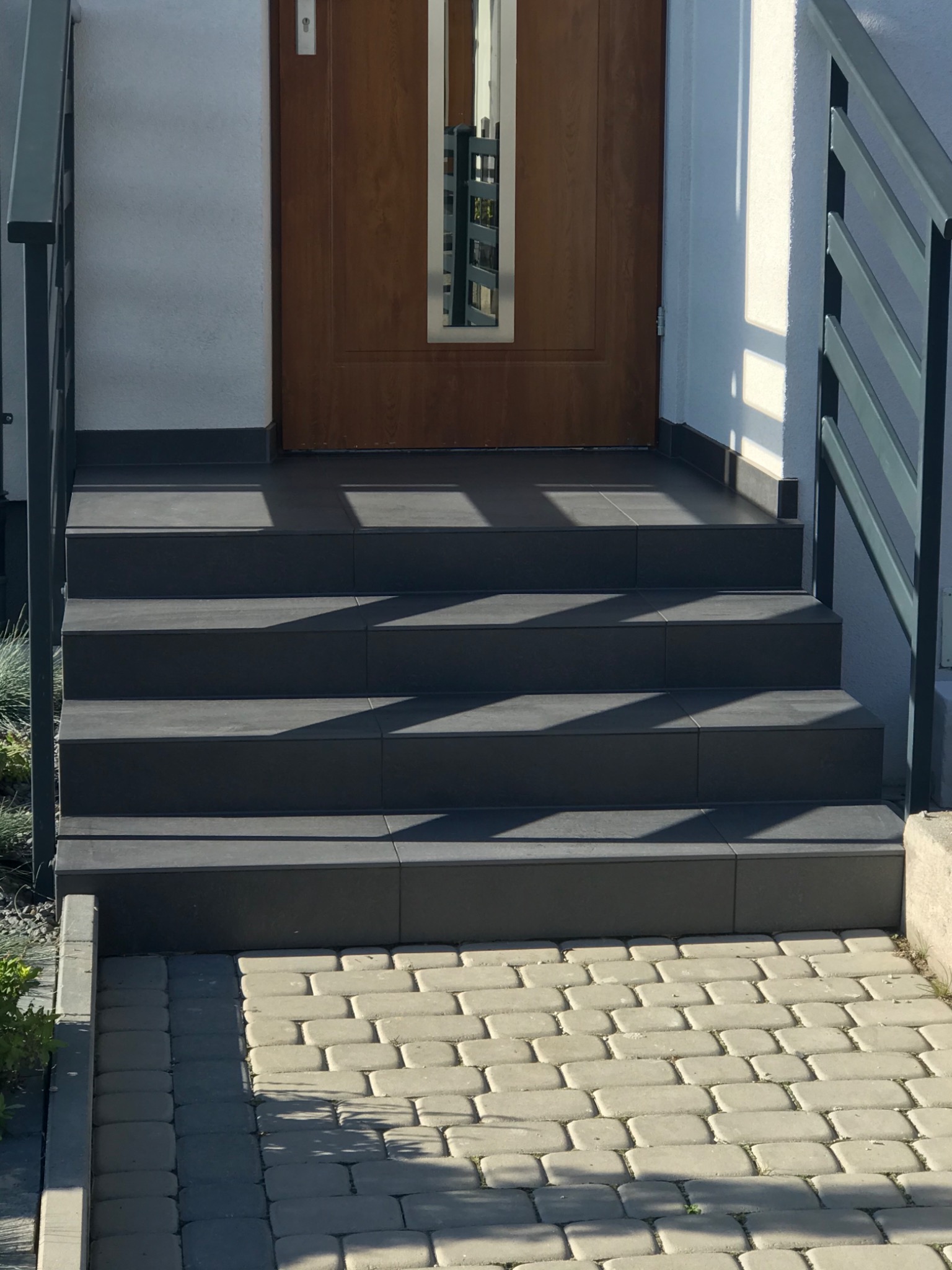 Zewnętrzne schody wejściowe wyłożone ciemnoszarą płytką, prowadzące do brązowych drzwi z przeszkleniem, z szarą metalową balustradą i brukowaną ścieżką.