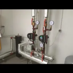 Złożona instalacja grzewcza z dwoma pompami obiegowymi, rurami pokrytymi izolacją termiczną i manometrami, zrealizowana w Pokoju. Widoczne zawory odcinające z czerwonymi uchwytami.