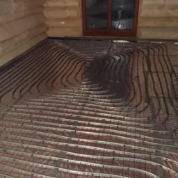 Instalacja ogrzewania podłogowego w drewnianym domu z bali, widoczne rury grzewcze ułożone na folii izolacyjnej z czerwoną siatką, przygotowane do zalania wylewką.