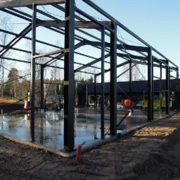Konstrukcja stalowa hali w Obornikach w trakcie budowy. Widoczne czarne słupy, stalowe belki i wylana posadzka. W tle drzewa i budynek.