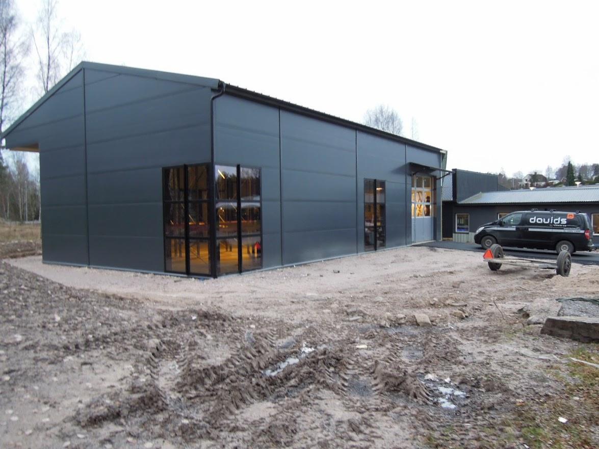 Szara hala z płyty warstwowej z dużymi oknami, widoczny fragment terenu przed budynkiem i samochód z przyczepą. Ujęcie z boku, w tle drzewa.