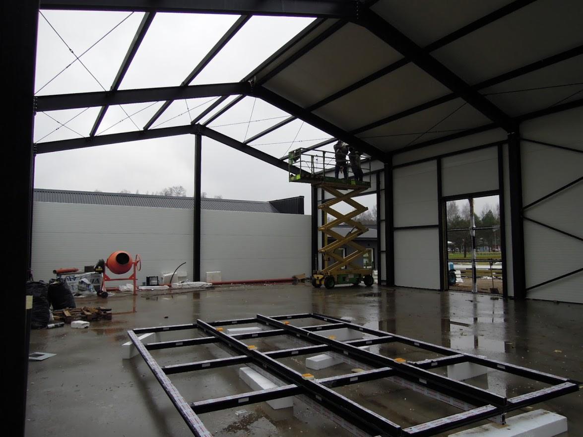 Wnętrze hali w budowie: stalowa konstrukcja dachu, pracownicy na podnośniku, ściany z płyt warstwowych, przygotowane ramy okienne na podłodze. Widoczny mikser do betonu.