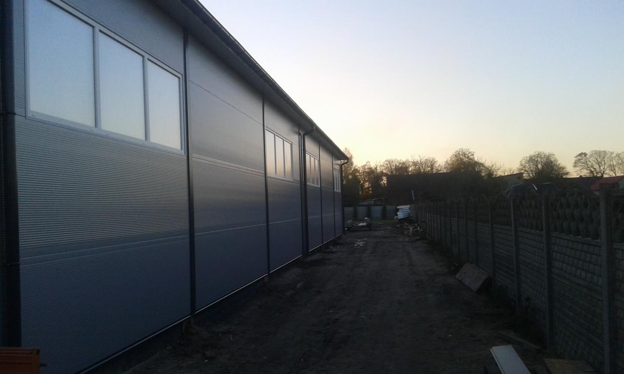 Szara hala z płyty warstwowej z oknami, widok z zewnątrz. Widoczne panele ścienne, okna i boczna ściana hali na tle wieczornego nieba i drzew.