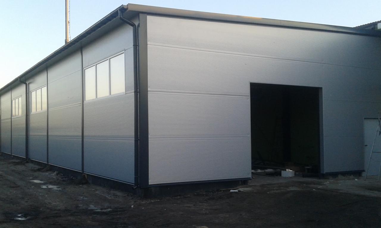 Zewnętrzny widok hali z szarej blachy trapezowej z oknami i otwartym wjazdem. Widoczny fragment rury wentylacyjnej na dachu. Ujęcie w pochmurny dzień.