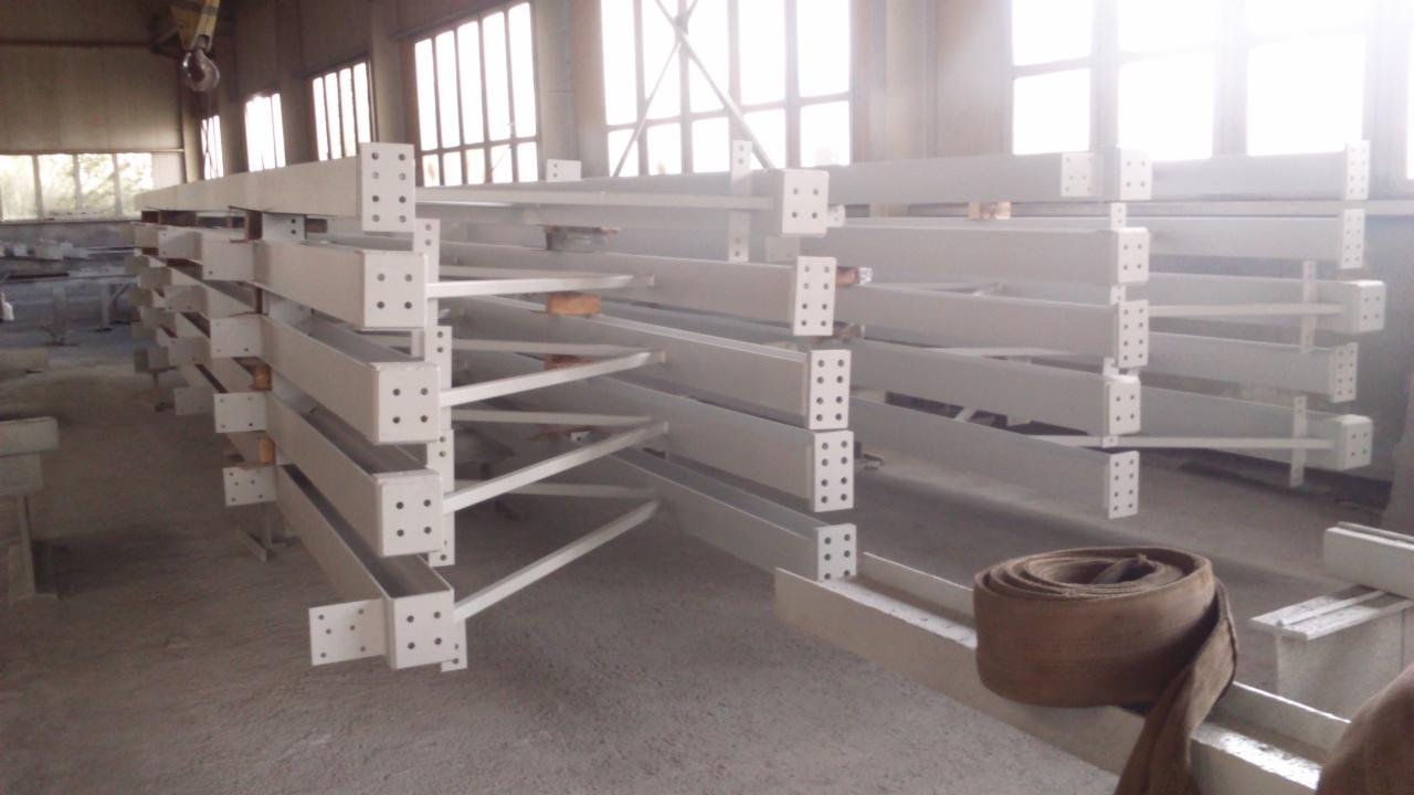 Białe, pomalowane elementy konstrukcji stalowej z otworami na śruby, ułożone warstwowo w hali, czekające na montaż. Widoczne okna i suwnica.