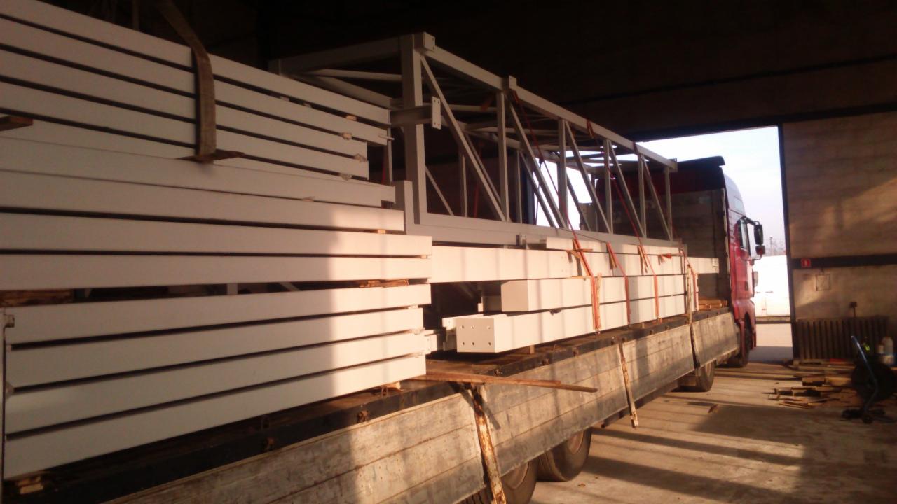 Ciężarówka z załadunkiem białych, stalowych elementów konstrukcyjnych, zabezpieczonych pasami, gotowych do transportu i montażu na placu budowy.