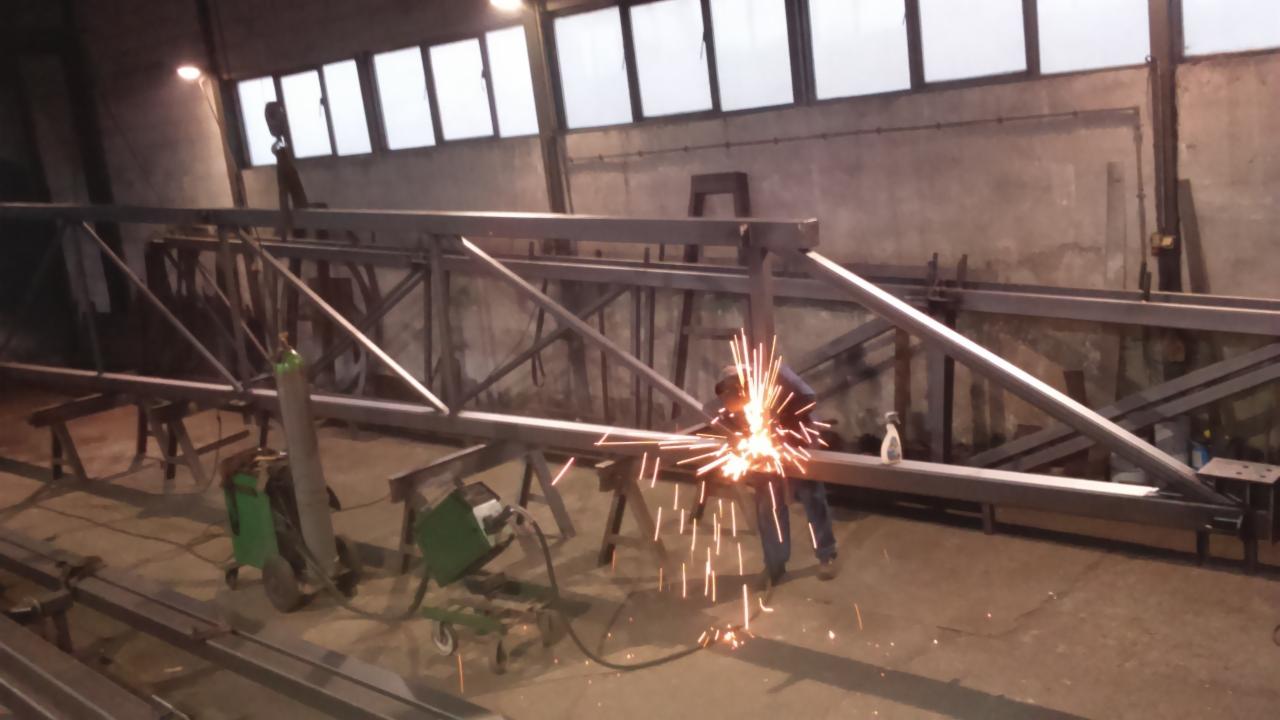 Spawanie konstrukcji stalowej w hali: spawacz łączy elementy, widoczne iskry, butla z gazem i spawarka. W tle okna i betonowa ściana.