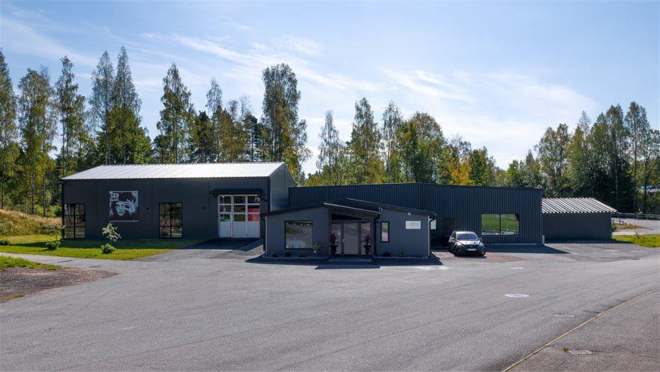 Nowoczesna, szara hala z biurem i parkingiem na tle zieleni. Budynek z blachy falistej i dużymi oknami. Na ścianie grafika.