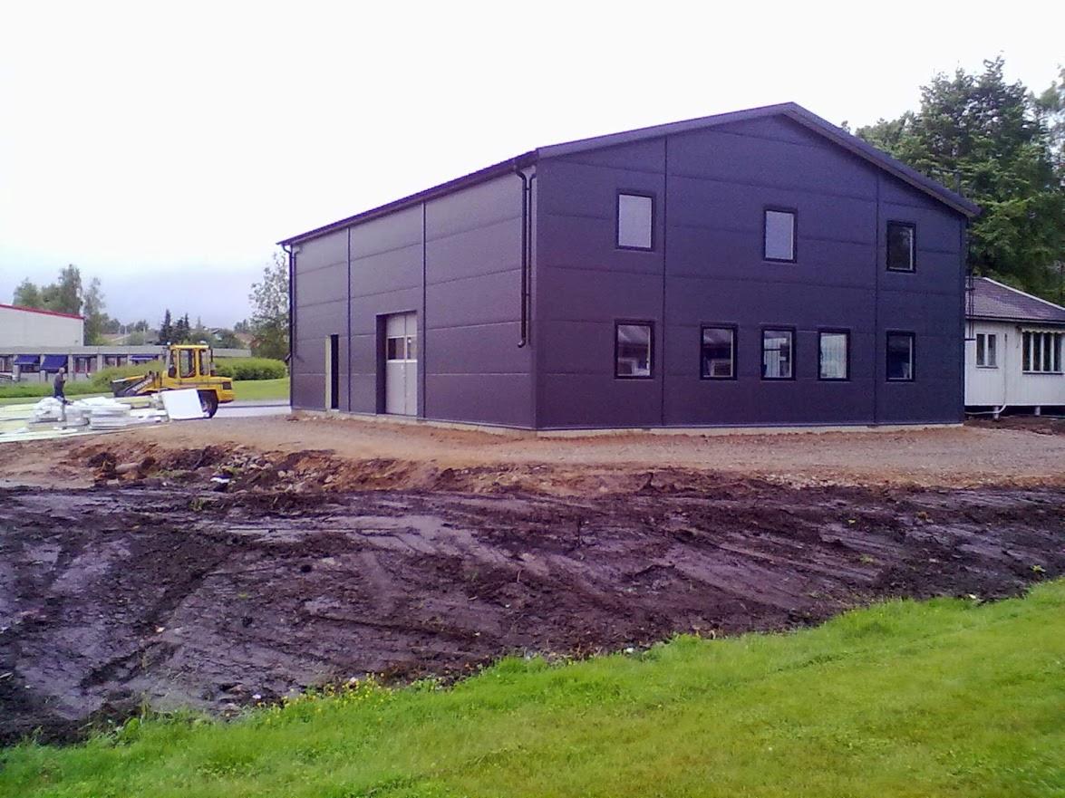 Nowoczesna hala z płyt warstwowych w grafitowym kolorze, z widocznymi oknami i drzwiami. W tle żółta koparka na terenie budowy.