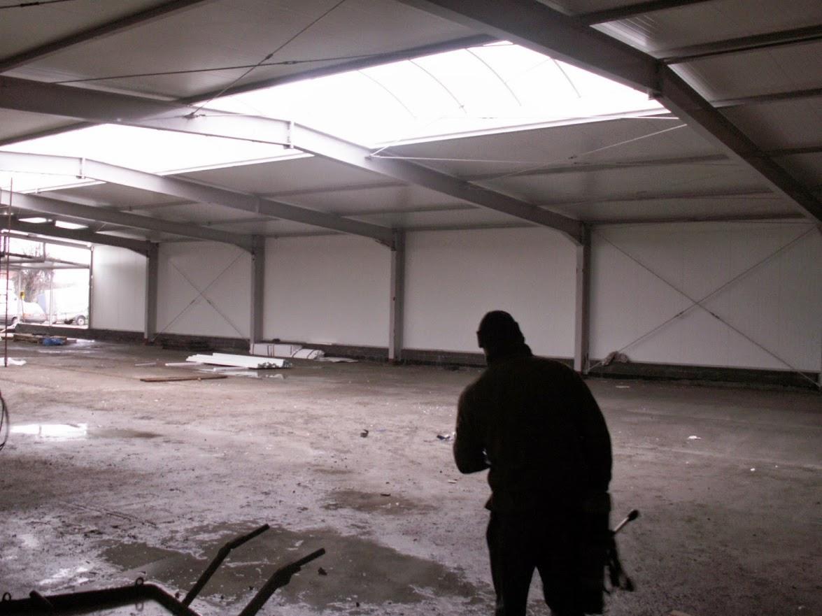 Wnętrze hali w trakcie budowy: widoczne stalowe konstrukcje, ściany z płyt warstwowych, świetlik dachowy i sylwetka osoby. Podłoże surowe, widać elementy rusztowania.