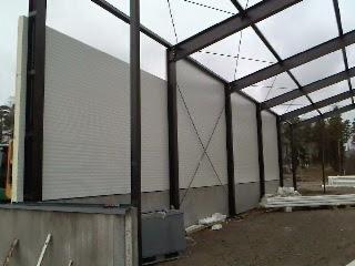 Wnętrze hali w budowie: stalowa konstrukcja i montaż białych płyt warstwowych. Widoczne elementy konstrukcyjne i fragment otoczenia zewnętrznego. Ujęcie z dołu.