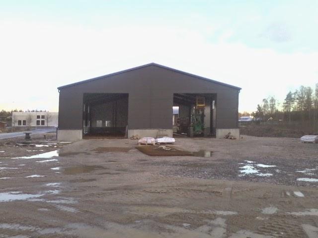 Szara hala stalowa z dwoma otwartymi wjazdami, w tle drzewa i budynek. Teren wokół hali jest błotnisty, co sugeruje niedawną budowę.