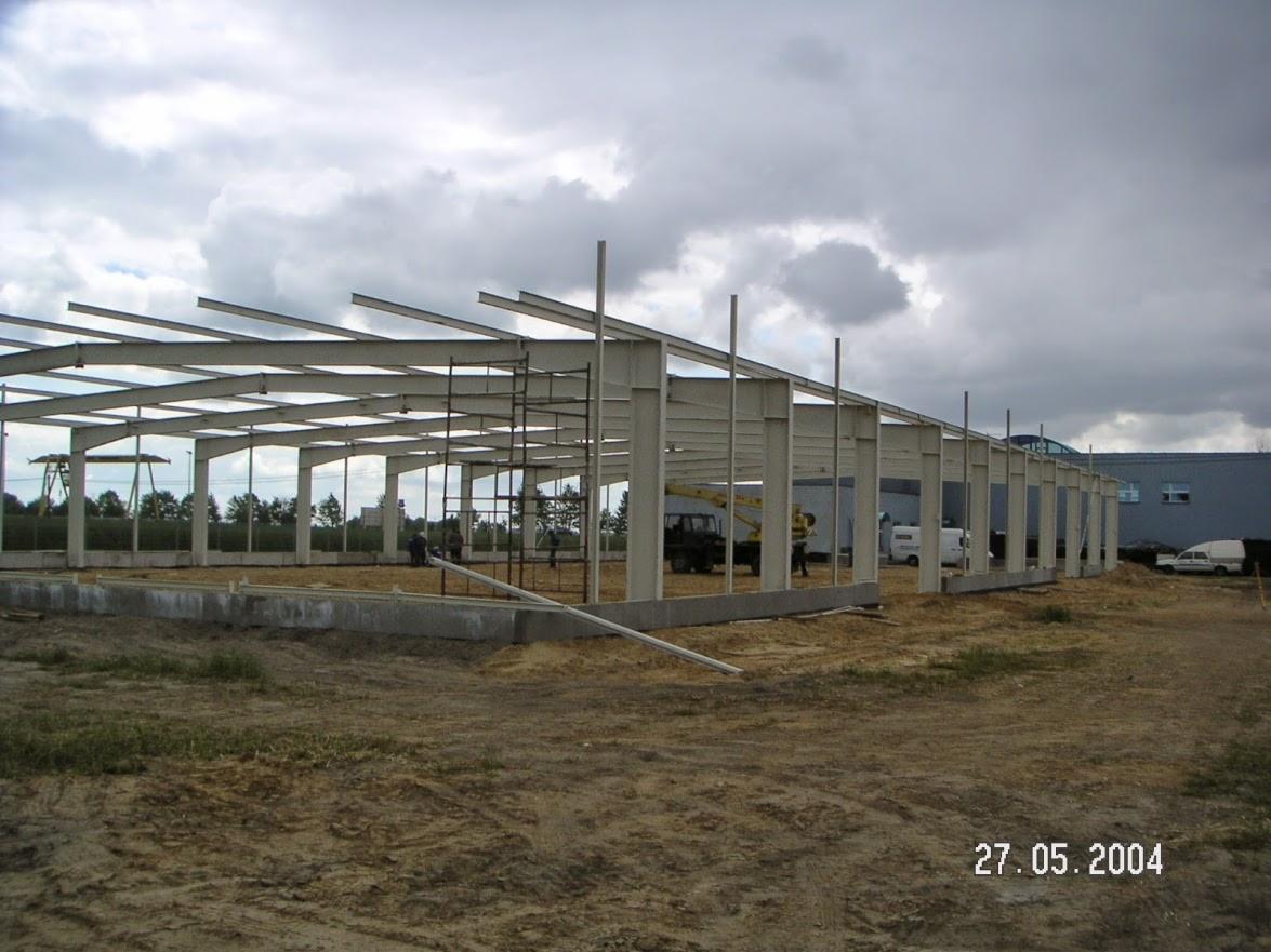 Szara stalowa konstrukcja hali w trakcie budowy, widok z zewnątrz. Na pierwszym planie podłoże, w tle budynek i pochmurne niebo. Ujęcie z dnia 27.05.2004.