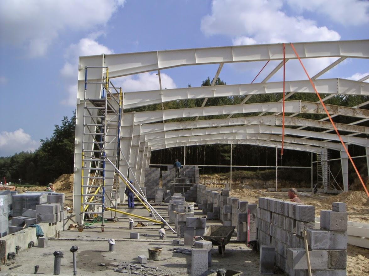 Budowa hali z białej konstrukcji stalowej i szarych bloczków betonowych w słoneczny dzień. Widoczne rusztowania i pracownicy.
