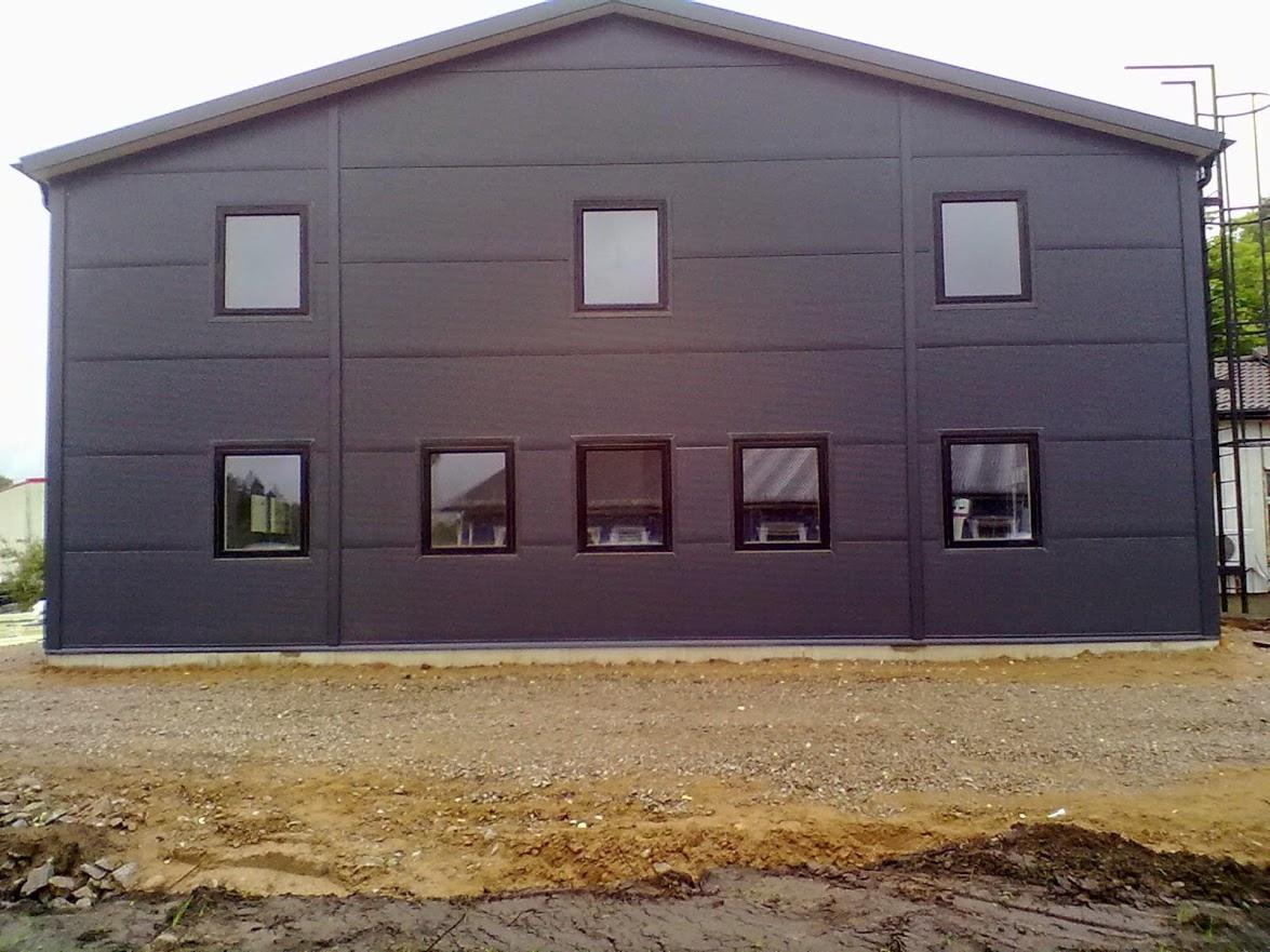 Nowoczesna hala z szarej płyty warstwowej z oknami, widok z zewnątrz. Na ziemi gruz i piasek. Widoczne łączenia płyt. Z prawej strony drabina.