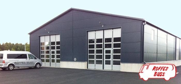 Szara hala z dwoma bramami segmentowymi z oknami, na tle błękitne niebo, obok zaparkowany bus z logotypem na boku. Widok z zewnątrz.