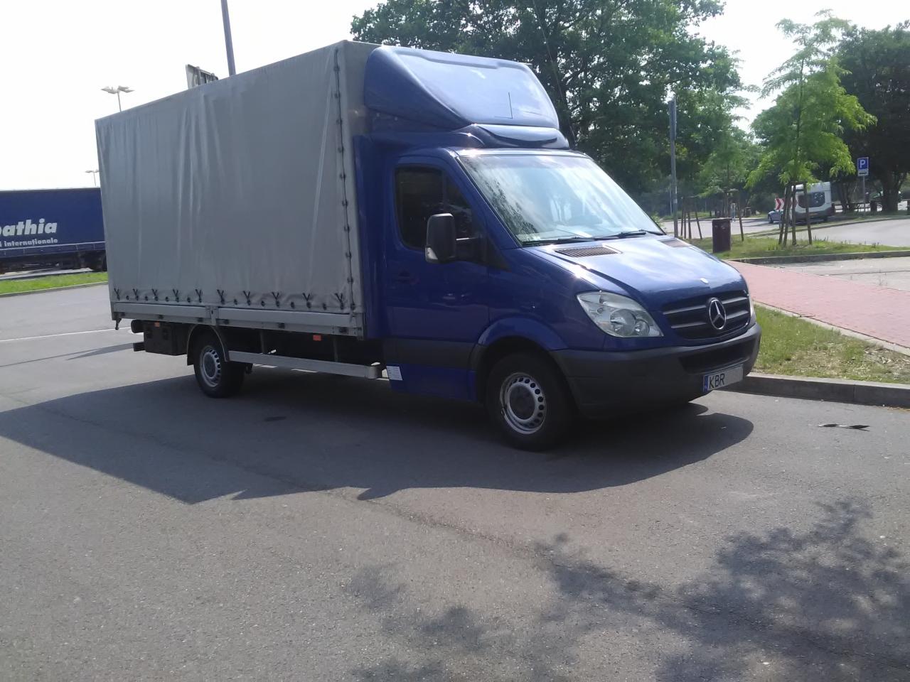 Niebieski samochód dostawczy Mercedes-Benz Sprinter z plandeką na parkingu, widoczny cień pojazdu, tablica rejestracyjna KBR.
