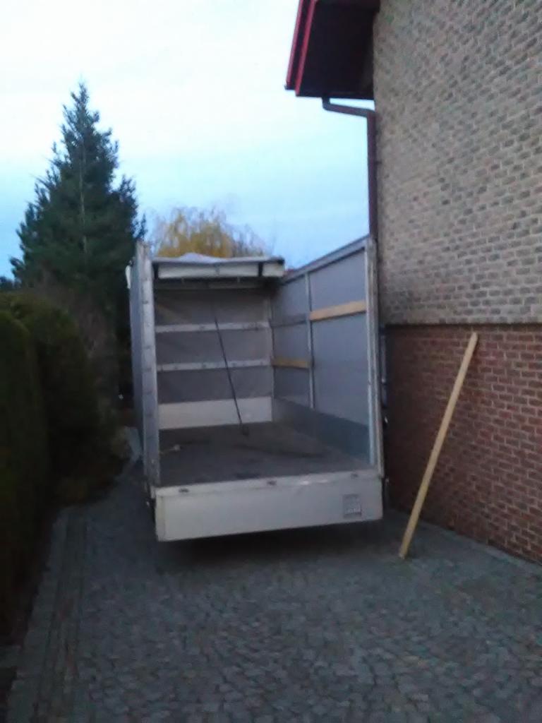Otwarta pusta paka samochodu dostawczego zaparkowanego tyłem do ceglanego budynku z drewnianą listwą opartą o ścianę, widoczny fragment drzewa iglastego w tle.