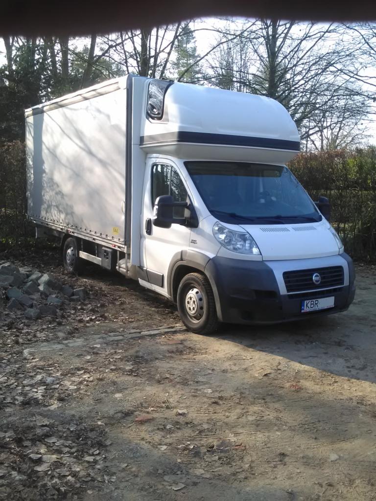 Biały samochód dostawczy Fiat z zabudową kontenerową, zaparkowany na grząskim terenie z liśćmi, widoczne drzewa w tle.