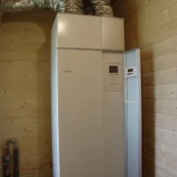 Biała pompa ciepła, z widocznymi rurami wentylacyjnymi, zamontowana w pomieszczeniu wykończonym drewnem. Detal instalacji wewnątrz budynku.